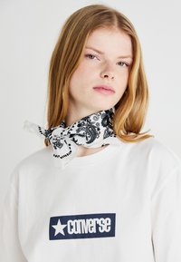 Weißes T-Shirt mit einem blauen "Converse"-Logo, kombiniert mit einem schwarz-weißen Bandana mit Paisleymuster um den Hals.