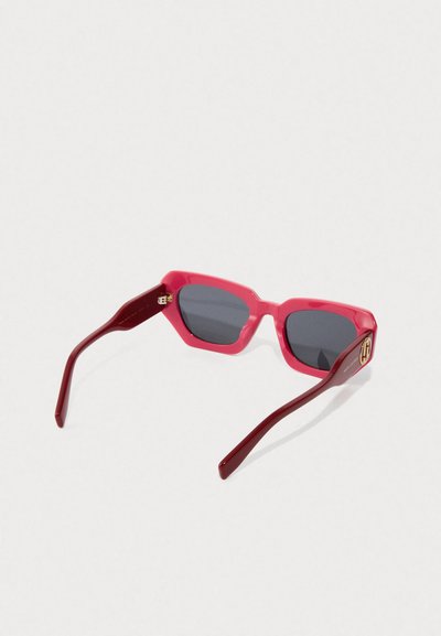 Rode rechthoekige zonnebril met donkere lenzen en een gouden embleem op de poot, weergegeven tegen een plain witte achtergrond.