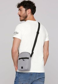 Graue Umhängetasche mit schwarzen Akzenten, rechteckige Form, strukturiertes Gewebe, mit einer vorderen Reißverschluss-Tasche und verstellbarem Riemen.