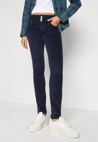 Mörkblå kordbyxors med smal passform och knäppning i midjan, framfickor, som bärs med vita platåskor. Accentuerad av en rutmönstrad skjorta.