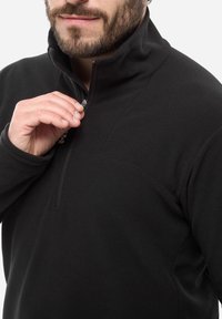 Schwarzer Fleece-Pullover mit hohem Kragen und halbseitigem Reißverschluss. Strukturstoff mit subtilen Mustern. Hand in der Nähe des Reißverschlusses positioniert.