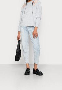 Sweat-shirt gris avec broderie florale orange sur les épaules, associé à un jean tapered bleu clair et des mocassins noirs épais. Sac noir inclus.