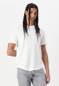 Weißes T-Shirt mit kurzen Ärmeln, lockerer Passform, strukturiertem Stoff, Rundhals und dezentem Design. Kombiniert mit hellgrauen Jeans.