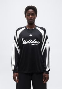 Άτομο που στέκεται φορώντας ένα μακρυμάνικο αθλητικό μπλουζάκι Adidas σε μαύρο και άσπρο χρώμα με λογότυπο και ρίγες στα μανίκια, μπροστά σε απλή φόντο.