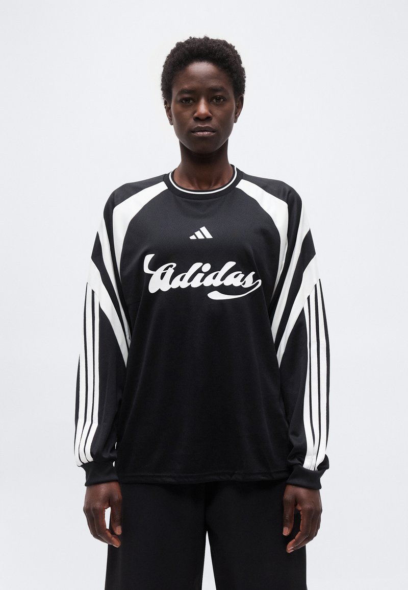 Άτομο που στέκεται φορώντας ένα μακρυμάνικο αθλητικό μπλουζάκι Adidas σε μαύρο και άσπρο χρώμα με λογότυπο και ρίγες στα μανίκια, μπροστά σε απλή φόντο.