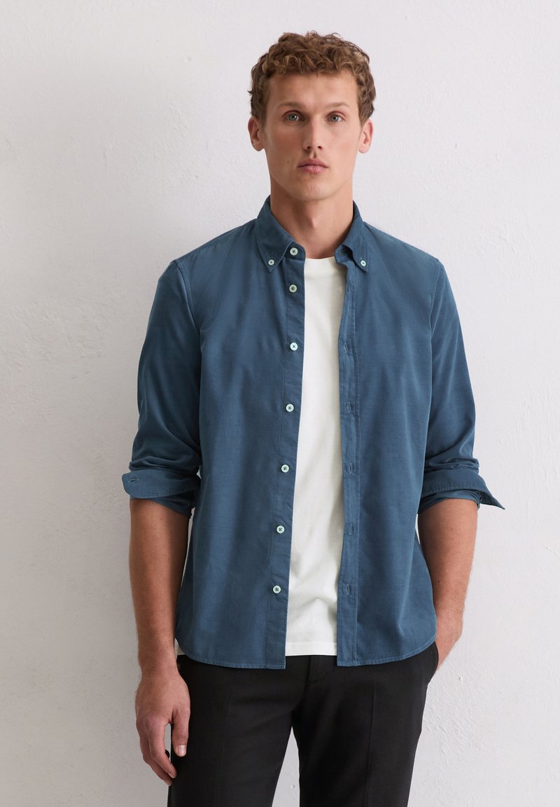 Dunkelblaues Hemd mit Knopfleiste, langärmlig mit hochgekrempelten Ärmeln, kombiniert mit einem weißen T-Shirt, minimalistisches Design, glatte Textur.