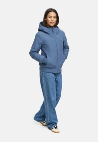 Veste à capuche bleue avec fermeture éclair à l'avant, poignets élastiques et deux poches latérales, associée à un jean bleu ample et des baskets décontractées.