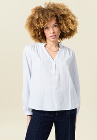 Blusa azzurro chiaro a maniche lunghe con righe bianche verticali, scollatura henley e colletto a volant. Tessuto di cotone, vestibilità rilassata.