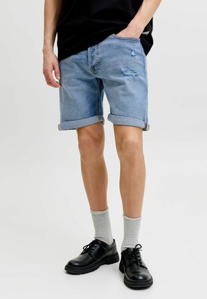 Osoba nosi svetlomodré vyhrnuté riflové šortky s miernym opotrebovaním, sivé ponožky, čierne šnurovacie topánky a čiernu košeľu.