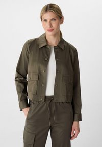 Olijfgroene cropped jack met knoopsluiting, twee voorzakken en een fijne textuur. Draagt bij een geribbelde witte top en bijpassende broek.