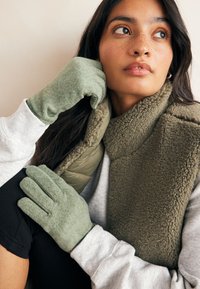 Groene fleecehandschoenen met een zachte textuur, gedragen bij een gezellige olijfgroene colbertjas en een lichtgrijs shirt met lange mouwen. Close-up weergave.