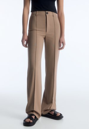 Pantaloni beige a vita alta con una vestibilità su misura, caratterizzati da una sola piega frontale, chiusura con bottone e un tessuto liscio e elasticizzato. Sandali neri.