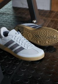 Witte sportschoenen met grijze strepen, glad bovenwerk van synthetisch materiaal, ronde neus en rubberen zool; geplaatst op een gestructureerde zwarte ondergrond.