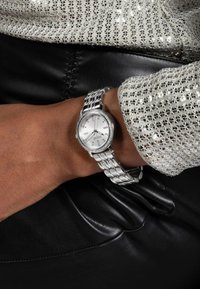 Silberne Uhr mit rundem Zifferblatt, strukturiertem Ziffernblatt, Metallgliederarmband und dekorativer Lünette, getragen an einem gebräunten Handgelenk, vor einem schwarzen Lederhintergrund.