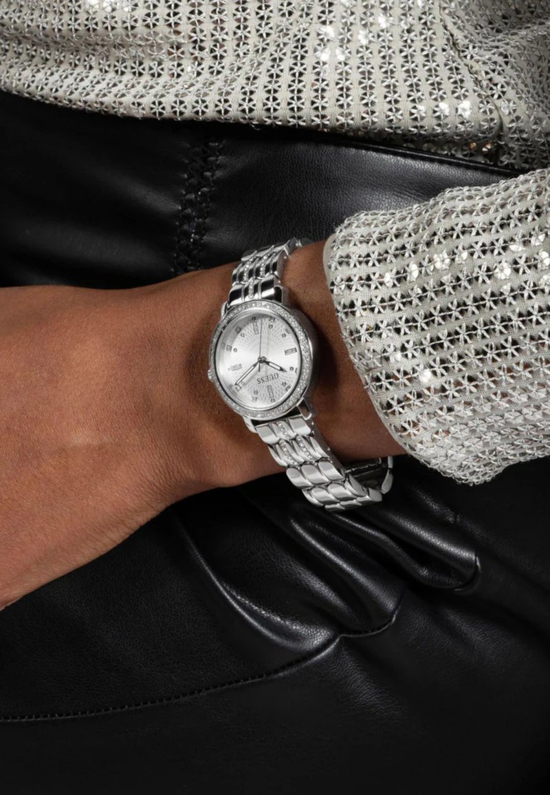 Silberne Uhr mit rundem Zifferblatt, strukturiertem Ziffernblatt, Metallgliederarmband und dekorativer Lünette, getragen an einem gebräunten Handgelenk, vor einem schwarzen Lederhintergrund.