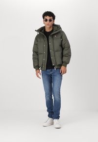 Chaqueta acolchada verde oliva con capucha, herrajes plateados, dos bolsillos frontales y detalle del logo. Combinada con jeans azules y zapatillas blancas.