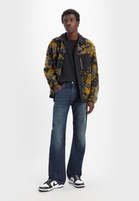 Multicolore fleece jack met een zwarte kraag en zak, gecombineerd met denim jeans met rechte pijpen en zwart-witte sneakers.