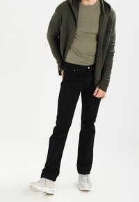 Olivgrön strukturerad hoodie som bärs över en åtsittande olivskjorta, ihop med svarta jeans och ljusgrå sportskor.