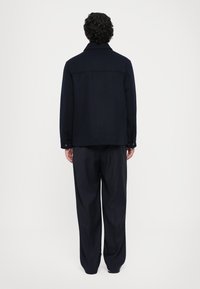 Mann med krøllete hår iført en mørk marineblå jakke og løse pinstripede bukser, står vendt mot en enkel hvit bakgrunn.