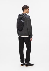 Borsa a tracolla nera con più scomparti con zip, indossata su un maglione grigio e pantaloni neri, realizzata in un materiale resistente e testurizzato.