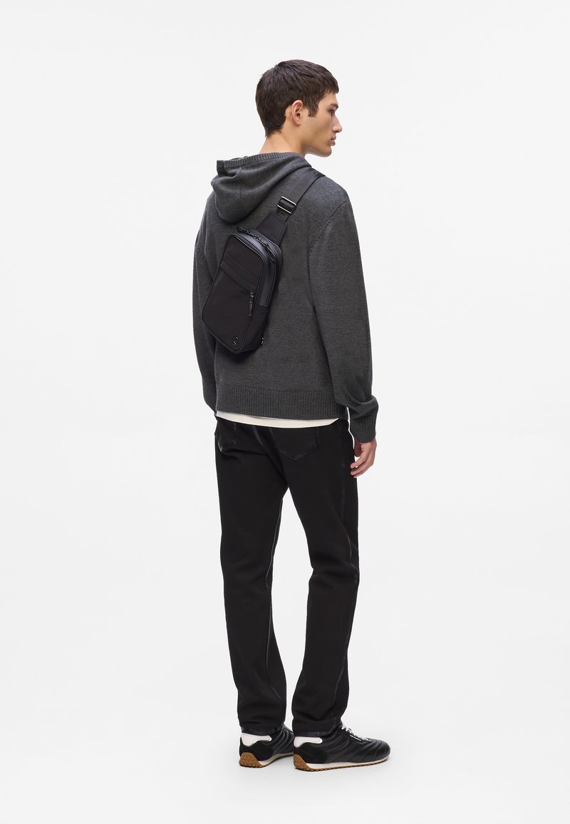 Borsa a tracolla nera con più scomparti con zip, indossata su un maglione grigio e pantaloni neri, realizzata in un materiale resistente e testurizzato.
