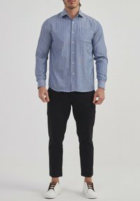 Chemise rayée bleu et blanc à manches longues avec un col boutonné, associée à un pantalon noir fuselé et des chaussures blanches avec des accents marron.
