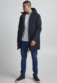 Mørkeblå parka-jakke med hette, med frontlommer, glidelås og svarte detaljer. Bært over en hvit skjorte og blå jeans.