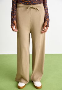 Pantaloni largi bej cu dungi, având un cordon la talie și deschideri laterale la tiv. Asortați cu adidași albi.