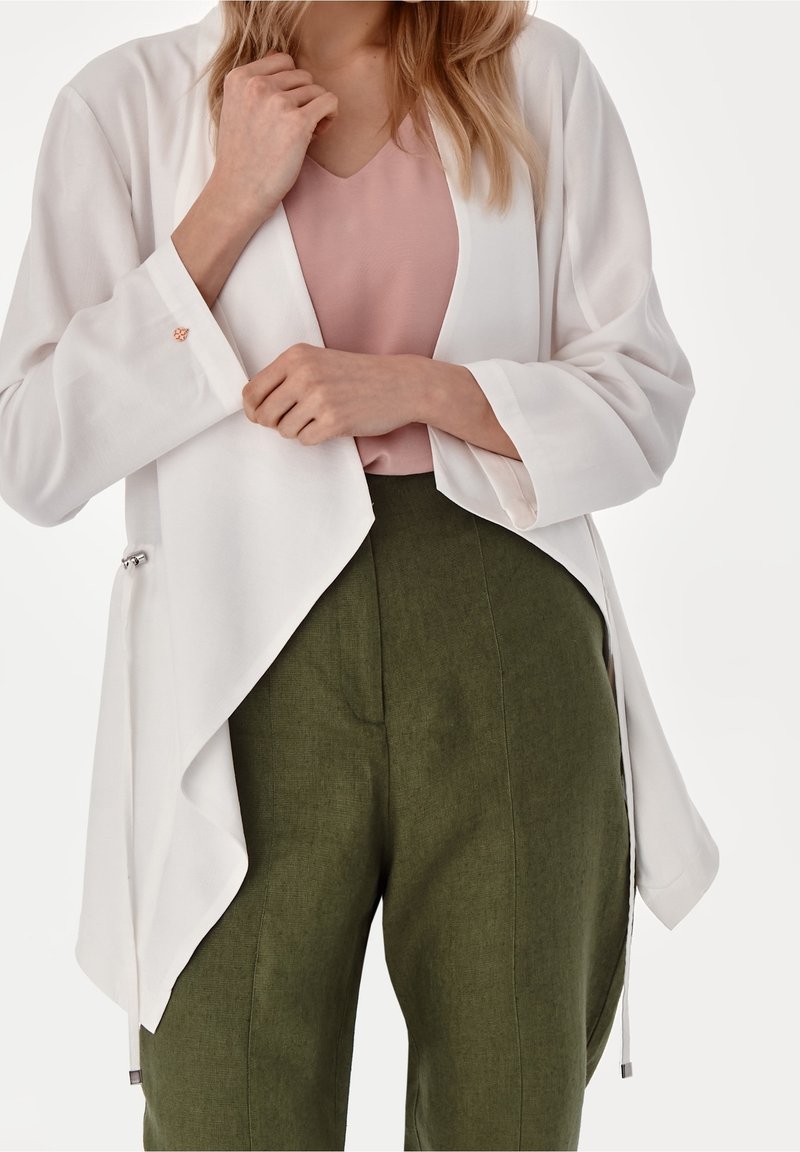 Femme portant une veste blanche ouverte sur un chemisier rose et un pantalon taille haute vert olive, ajustant la veste avec ses mains.