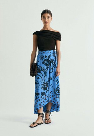 Mujer que lleva un top negro sin hombros, falda cruzada azul con flores, sandalias negras con cordones, sostiene un bolso de mano negro con abalorios, de pie frente a un fondo blanco.