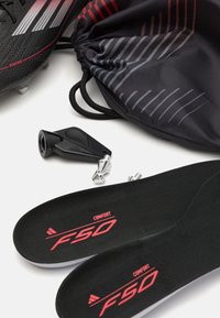 Black soccer cleats with red accents, a black drawstring bag with gray stripes, black insoles labeled "COMFORT F50," and black and silver hardware.