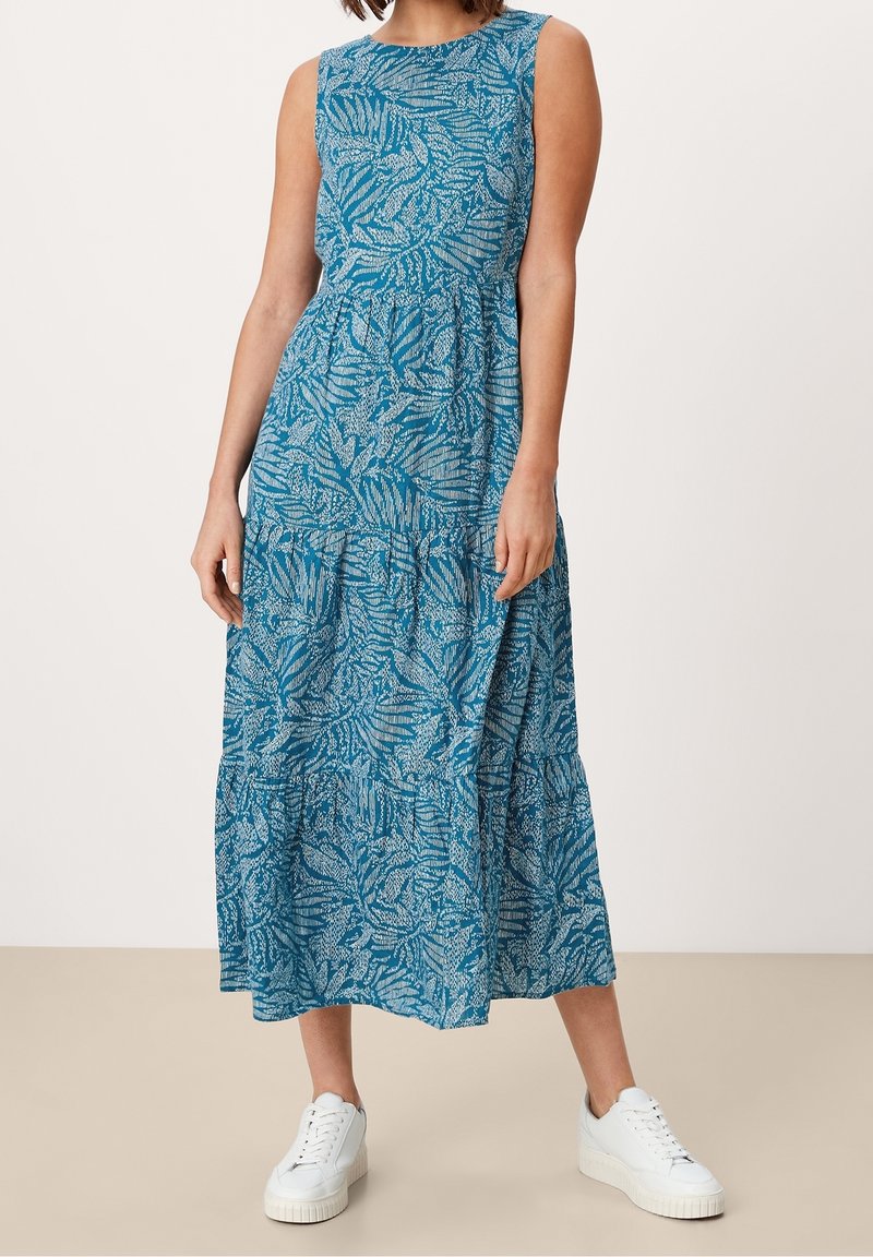 Une robe maxi bleue avec un motif de feuilles, présentant un col rond, un design sans manches et une jupe à volants. Associée à des baskets blanches.