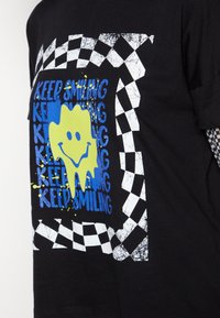 T-shirt in cotone nero con un design grafico blu e verde featuring una faccina sorridente e del testo ripetitivo che dice "CONTINUA A SORRIDERE", bordato da un motivo a scacchi.