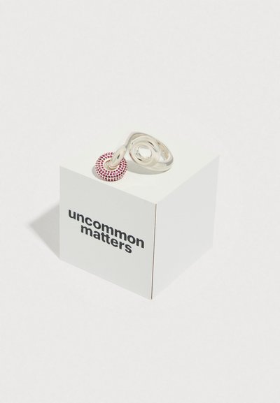 Bague en argent présentant des spirales entrelacées, agrémentée d'une pierre rouge circulaire incrustée de petits cailloux, exposée sur une boîte blanche.