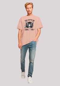 Light pink cotton t-shirt featuring a graphic of a cat in sunglasses with text saying "You Gotta Be Kitten Me!" Paired with distressed blue jeans and white sneakers.
