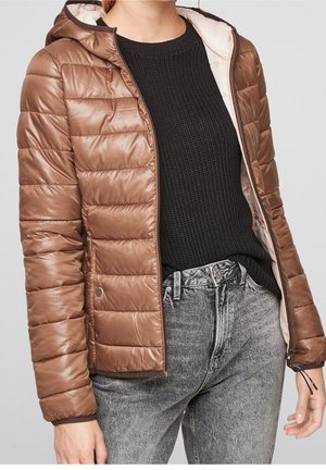 Personne portant une veste matelassée à capuche marron par-dessus un pull en maille noir et un jean gris délavé sur un fond uni.