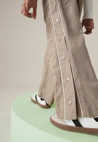 Pantalons beige à large jambe avec des boutons décoratifs et des accents blancs, associés à des baskets blanches et noires sur un piédestal vert.