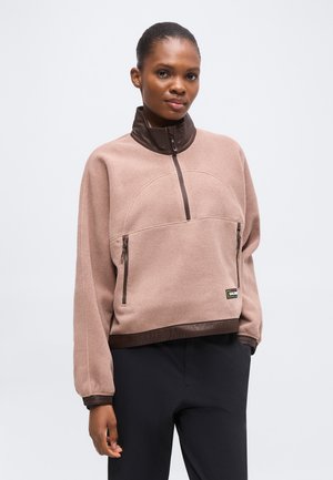 Kvinde iført beige fleecejakke med lynlås og brun kant samt sorte bukser, stående foran en ensfarvet lys baggrund.