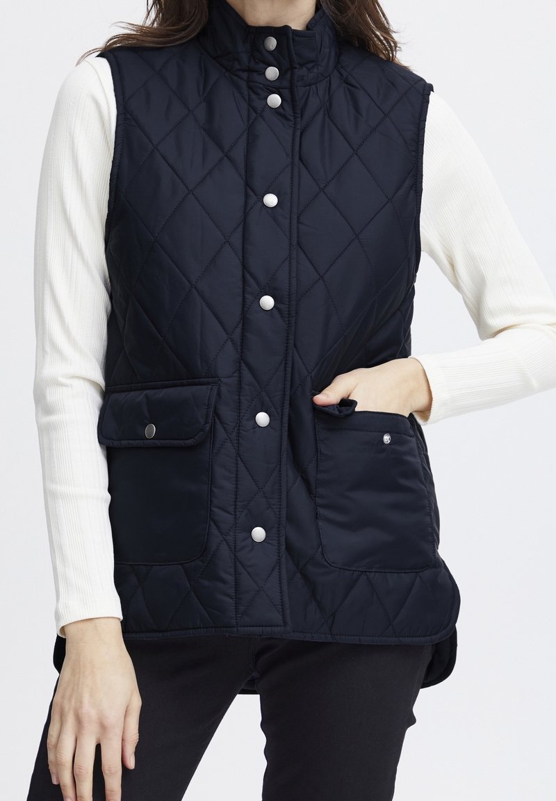 Femme portant une veste matelassée sans manches bleu marine avec des boutons argentés et deux poches avant à rabat, par-dessus un haut blanc à manches longues et un pantalon noir.