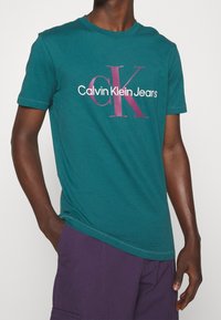 Tyrkysové bavlněné tričko s bílým a vínovým logem "Calvin Klein Jeans", kulatým výstřihem a krátkými rukávy.