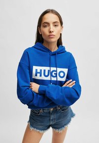 Blue hooded sweatshirt with white "HUGO" logo, soft fabric, loose fit, and ribbed cuffs. Paired with denim shorts featuring frayed edges.
