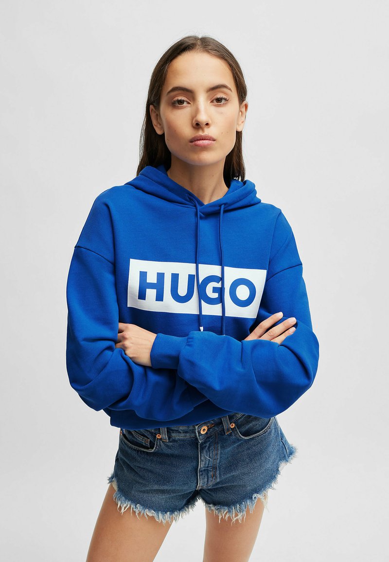Blue hooded sweatshirt with white "HUGO" logo, soft fabric, loose fit, and ribbed cuffs. Paired with denim shorts featuring frayed edges.