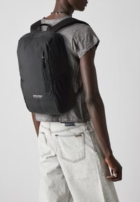 Zaino nero in nylon con tasca frontale con zip, spallacci imbottiti e design rettangolare semplice. Indossato sopra una maglietta grigia e jeans chiari.