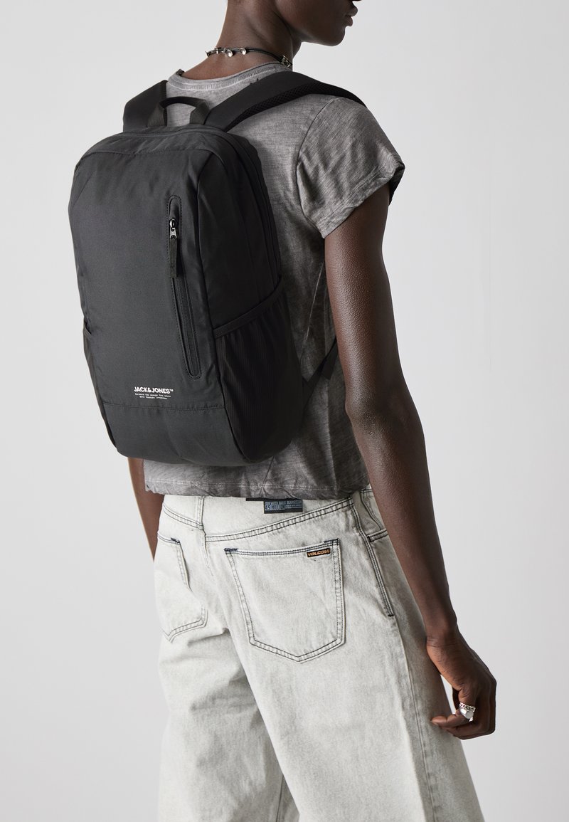Zaino nero in nylon con tasca frontale con zip, spallacci imbottiti e design rettangolare semplice. Indossato sopra una maglietta grigia e jeans chiari.