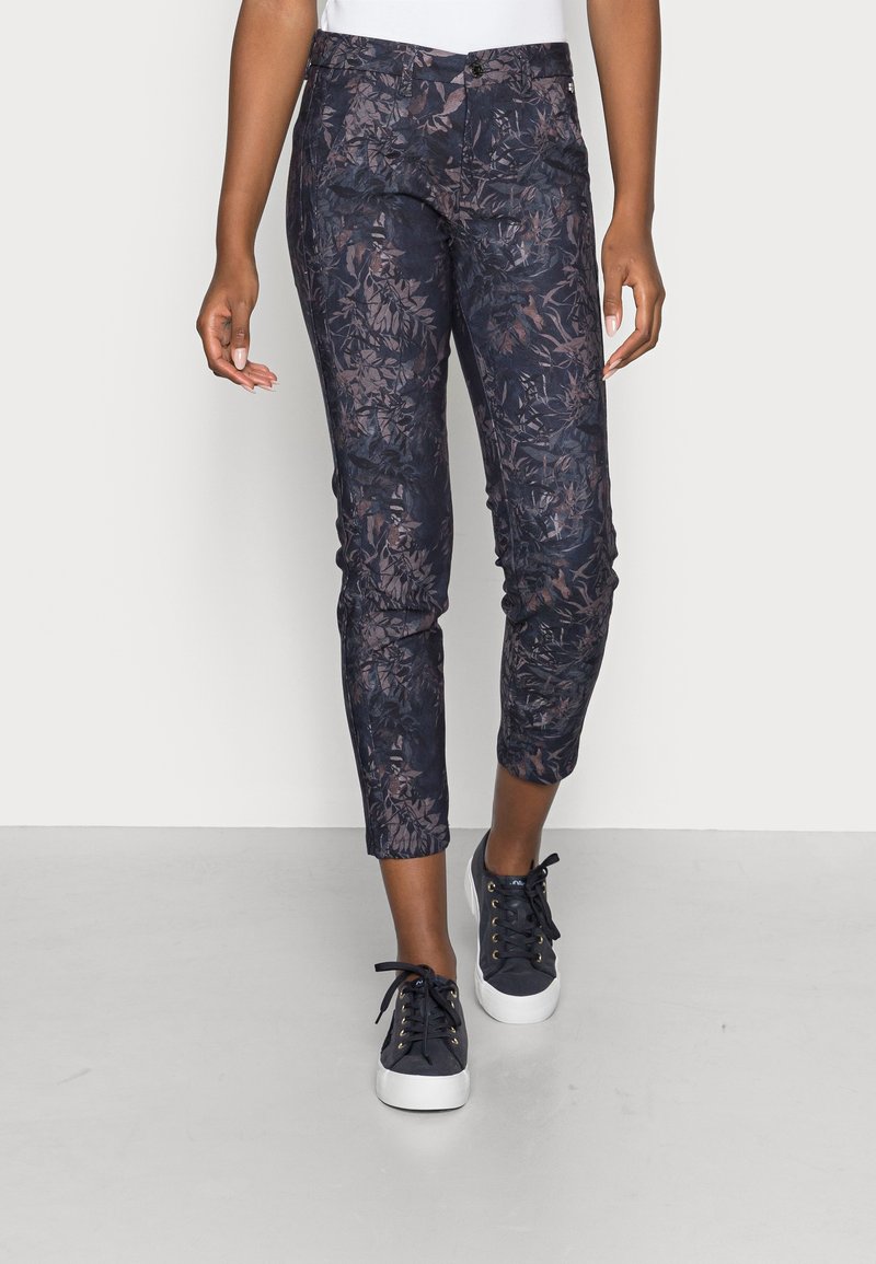 Pantalon slim raccourci avec motif floral sombre porté avec des baskets noires à lacets sur une personne debout sur fond blanc.