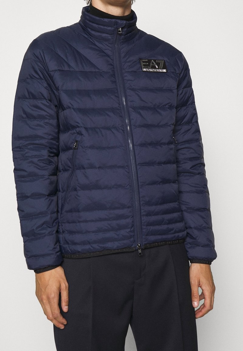 Marinblå puffer-jacka med vertikal quiltad design, hög krage, dragkedjestängning och EA7-logotyp på bröstet. Har sidofickor och elastisk nederkant.