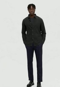 Άνδρας στέκεται με τα χέρια στις τσέπες, φορώντας μαύρο πουκάμισο με κουμπιά, σκούρα μπλε παντελόνι και μαύρα παπούτσια, μπροστά σε έναν απλό γκρι φόντο.