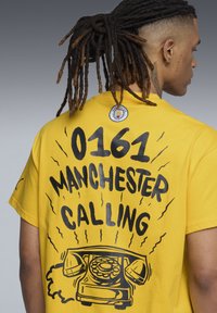 Gul bomulds t-shirt med fed sort tekst "0161 MANCHESTER CALLING" og et grafisk motiv af en vintage telefon på ryggen.