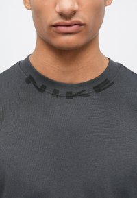 Camiseta de algodón gris con cuello redondo. Presenta un sutil logo negro de NIKE en el cuello. Textura suave, ajuste regular.