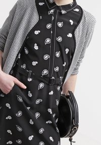 Schwarzes Kleid mit weißen Blumen- und Paisleymustern, kombiniert mit einer grauen gemusterten Strickjacke, die über einen Kragen und einen Gürtel verfügt. Schwarze Handtasche inklusive.